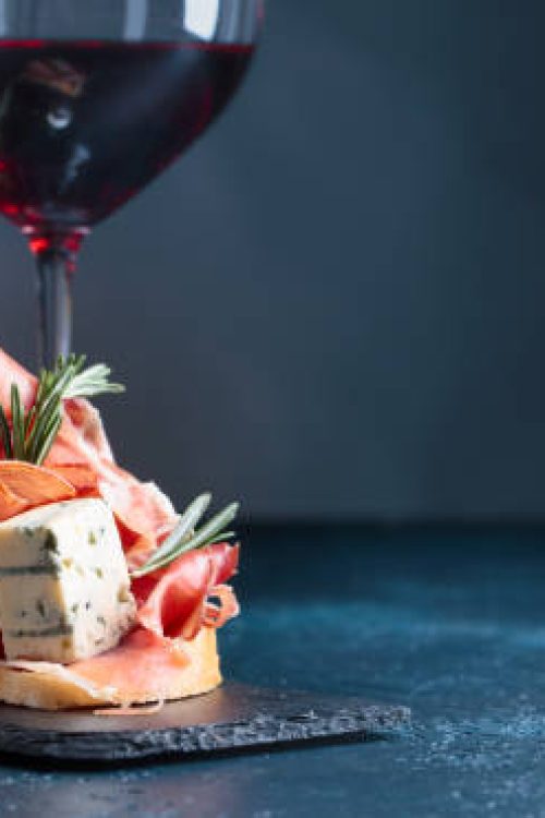 Sandwich with prosciutto, blue cheese and rosemary on a dark background. Glasses and bottle of red wine with snack. Copy space for your text.