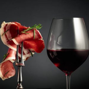 Glass of red wine and sliced prosciutto with rosemary on a fork, black background.